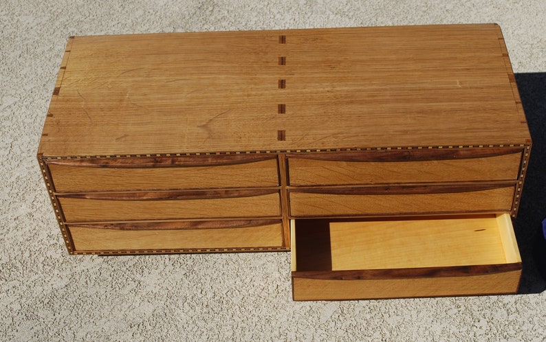 May include: A wooden chest of drawers with six drawers, one partially open. The chest is made of light brown wood with darker brown accents and decorative inlays. The drawers have curved handles and the chest is rectangular.