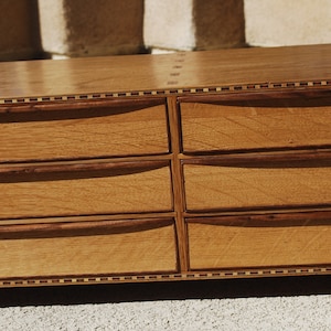 May include: A six-drawer wooden dresser with a natural finish. The drawers have curved handles and are framed by darker wood trim. The top and edges feature a decorative inlaid border. The dresser sits on short, angled legs.