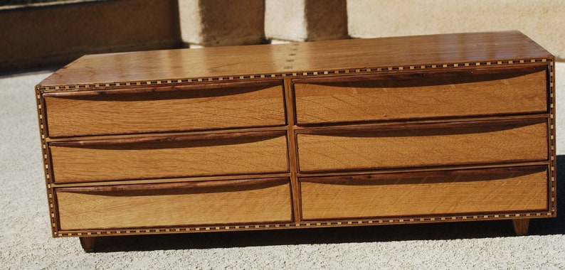 May include: A six-drawer wooden dresser with a natural finish. The drawers have curved handles and are framed by darker wood trim. The top and edges feature a decorative inlaid border. The dresser sits on short, angled legs.