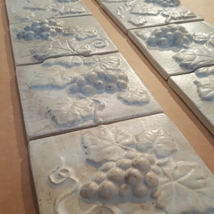 May include: A set of white ceramic tiles with a raised design of grapes and leaves. The tiles are arranged in rows and are perfect for a kitchen backsplash or bathroom wall.