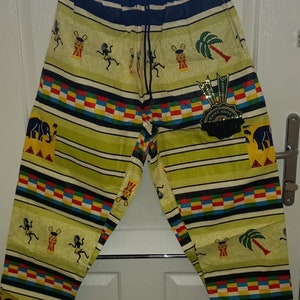 Puede incluir: Pantalones de patrón amarillo y negro con un diseño de rayas de colores. Los pantalones tienen una cintura con cordón y presentan gráficos de elefante, palmera y tambor.
