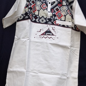 Peut inclure: Chemise à manches courtes blanche avec un motif géométrique noir, blanc et rouge sur la moitié supérieure. La chemise a une fermeture à bouton unique au niveau du cou et une petite poche sur le côté gauche. La moitié inférieure de la chemise présente un triangle blanc avec un contour brun et le mot "Wunu" brodé en brun.