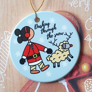 May include: A round Christmas ornament with a light blue background featuring a cartoon figure in a red coat and a reindeer. The ornament has the text "Dashing through the snow." A gold string is attached for hanging.