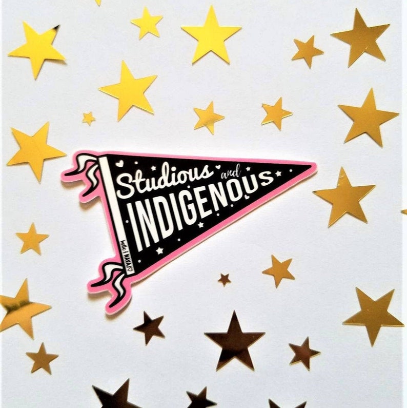 May include: A sticker in the shape of a pennant with the words "Studious and Indigenous" in white on a black background, outlined in pink. The sticker is surrounded by gold and yellow star-shaped cutouts on a white background.