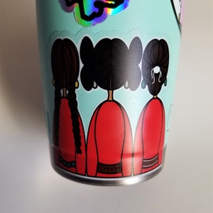 May include: A light teal tumbler adorned with stickers. One sticker features a holographic design, while another depicts three figures in red dresses with black hair. The tumbler has a silver base.
