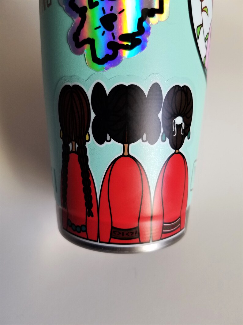 May include: A light teal tumbler adorned with stickers. One sticker features a holographic design, while another depicts three figures in red dresses with black hair. The tumbler has a silver base.
