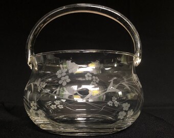 Könnte beinhalten: Eine klare Glasschüssel mit Henkel, verziert mit einem geätzten Blumenmuster. Die Schüssel hat eine abgerundete Form und eine breite Öffnung.