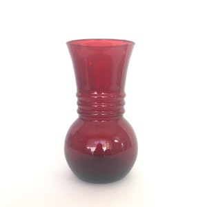 May include: A ruby red glass vase with a bulbous base and a flared top. The vase has a series of horizontal ridges around the middle. The glass is transparent, allowing light to pass through and reflect off the surface.