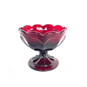 May include: A ruby red glass pedestal bowl with a scalloped rim. The bowl has a rounded base and a stem. The glass has a glossy finish and is likely vintage. The bowl is designed for serving desserts or displaying small items.