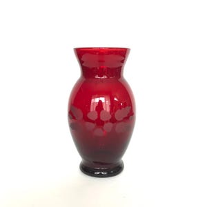 Puede incluir: Un jarrón de cristal rojo rubí con cuerpo redondeado y abertura acampanada. El jarrón presenta una base oscura y sutiles diseños florales de color más claro. La superficie lisa y brillante refleja la luz, resaltando su forma elegante.