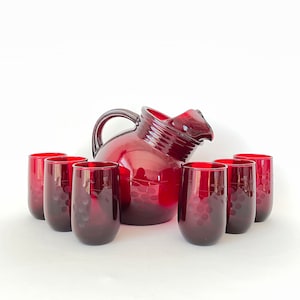 Puede incluir: Un juego de seis vasos rojos de vidrio y una jarra roja de vidrio con un diseño de uva. La jarra tiene un asa y una boca ancha.