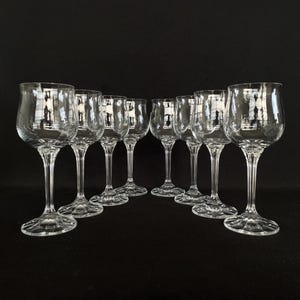 May include: Set of eight clear glass wine glasses with a rounded bowl and a long stem. The glasses are arranged in a row on a black surface.