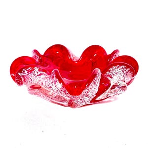 May include: A bright red glass ashtray with a ruffled, petal-like design. The edges have a speckled, white pattern, adding texture to the glossy, transparent bowl. This decorative piece is ideal for home decor.