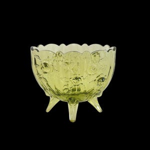 May include: A light green glass bowl with a scalloped edge, standing on three legs. The bowl has a floral design and is set against a black background. The glass has a translucent quality. The bowl's diameter is approximately 15 cm.
