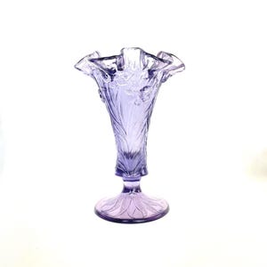 May include: A lavender-colored glass vase with a ruffled rim and a flared base. The vase features an etched floral design on the body and base. The vase is approximately 10 inches tall.