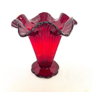 May include: A vibrant ruby red glass vase, featuring a ruffled rim and a fluted body. The vase has a scalloped base and a flared, trumpet-like shape. The glass is transparent, allowing light to pass through.