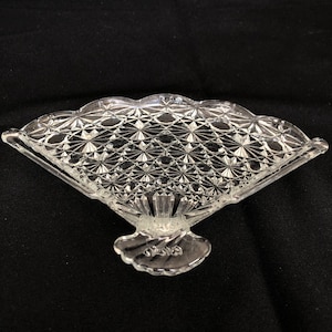 Könnte beinhalten: Eine klare, fächerförmige Schale aus Glas mit einem dekorativen Diamantenmuster. Die Schale hat einen gewellten Rand und einen Sockel.
