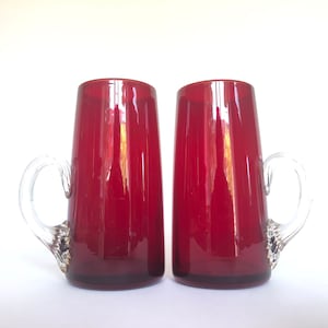 Könnte beinhalten: Zwei rote Glasbecher mit klaren Griffen. Die Becher sind hoch und zylindrisch mit einem abgerundeten Boden.