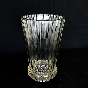 Könnte beinhalten: Eine transparente Glasvase mit vertikalen Rippen und einem strukturierten, abgerundeten Boden. Die Vase hat eine ausgestellte Form und ist für Blumen oder andere dekorative Gegenstände konzipiert. Das Glas ist transparent.
