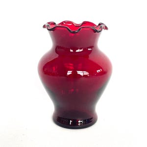 May include: A deep red glass vase with a frilled rim. The vase has a rounded body and a flared base. The glass is smooth and reflective, with a glossy finish. The vase is a decorative item.