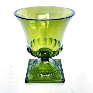 Op de afbeelding: Een groene glazen vaas met een vierkante basis en een uitlopende bovenkant. De vaas heeft een decoratief ontwerp rond de basis van de kom en op de vierkante basis. Het glas heeft een kleine scheur bij de rand.