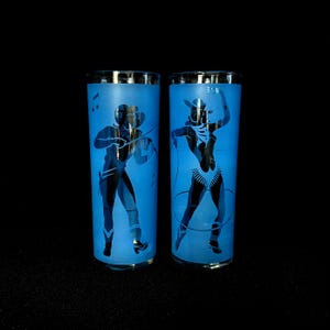 May include: Two tall, blue-tinted drinking glasses with silver rims. Each glass features a frosted silhouette design of a person. One glass depicts a musician, the other a cowgirl. The background is black.