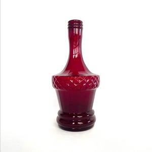 May include: A deep red glass bottle with a diamond pattern around the middle. The bottle has a long, slender neck and a rounded base with two circular ridges. The bottle is empty and is set against a white background.