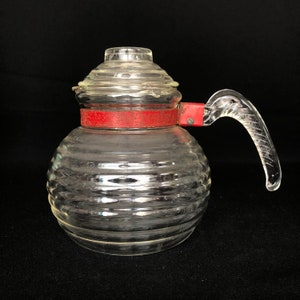 May include: A clear glass teapot with a red metal band around the top and a clear glass handle. The teapot has a ribbed design.