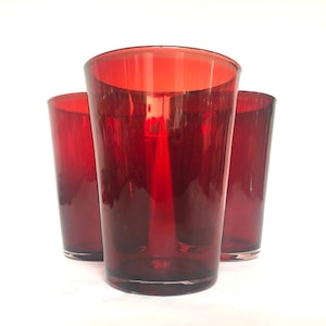 May include: Three red drinking glasses. The glasses are made of a transparent material and have a deep red colour. The glasses are cylindrical with a slightly flared top. The glasses are arranged in a group, with one in front and two behind.