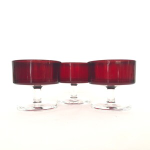 Könnte beinhalten: Drei rote Glasschalen für Desserts mit klaren Glasstielen und -füßen. Die Schalen sind in einer Reihe angeordnet.