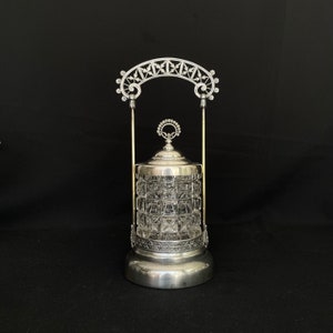 Pode incluir: Um pote de prata e vidro com uma tampa decorativa e uma alça de prata em forma de arco. O pote tem um corpo de vidro transparente com um padrão geométrico.