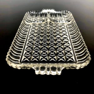 May include: A clear, rectangular glass serving tray with a geometric pattern. The tray has a scalloped edge and two small handles. The center features a repeating diamond design, adding a touch of elegance to the serving piece.