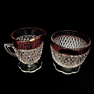 May include: A clear glass creamer and sugar bowl set with a diamond pattern and a red rim. The creamer has a handle and a spout. The sugar bowl is round. Both pieces sit on a clear, scalloped base.