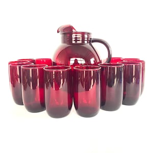 Puede incluir: Un juego de cristalería vintage de color rojo rubí. El juego incluye una jarra grande con asa y boquilla, rodeada por una docena de vasos altos a juego. La cristalería es de un rojo profundo y transparente.