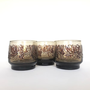 Puede incluir: Tres vasos de vidrio marrón con un diseño de hojas doradas. Los vasos son transparentes y tienen una forma redondeada.