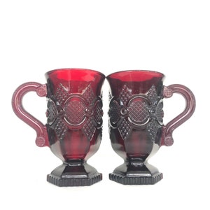 Op de afbeelding: Twee rode glazen bekers met een decoratief patroon. De bekers hebben een handvat en een brede basis.