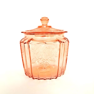Könnte beinhalten: Ein Vintage-Glasbehälter in Rosa mit Deckel. Der Behälter hat ein geätztes Blumendesign auf der Vorderseite und ist achteckig geformt. Der Deckel hat einen kleinen Knopf. Der Behälter dient wahrscheinlich zur Aufbewahrung.