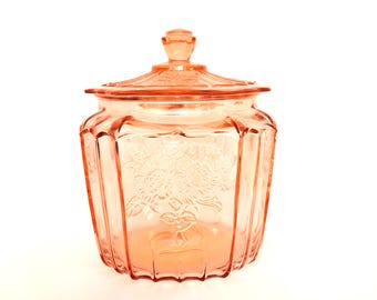 Pode incluir: Um frasco de vidro rosa vintage com tampa. O frasco tem um desenho floral gravado na frente e tem formato octogonal. A tampa tem um pequeno botão. O frasco é provavelmente usado para armazenamento.