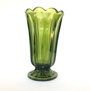 Puede incluir: Un jarr&oacute;n de vidrio verde con un borde festoneado y una base de pedestal. El jarr&oacute;n tiene detalles verticales acanalados y una forma acampanada. El color es un verde oliva apagado, y el vidrio parece ser vintage.