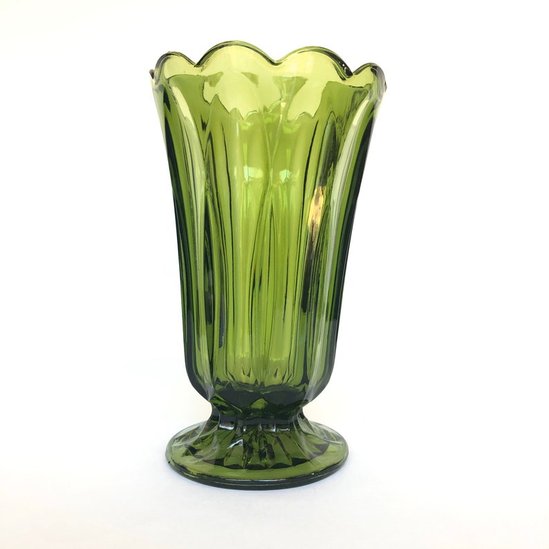Puede incluir: Un jarr&oacute;n de vidrio verde con un borde festoneado y una base de pedestal. El jarr&oacute;n tiene detalles verticales acanalados y una forma acampanada. El color es un verde oliva apagado, y el vidrio parece ser vintage.