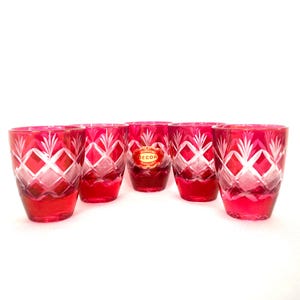 May include: Six ruby red drinking glasses with a cut crystal design. The glasses feature a diamond and leaf pattern. A red and gold sticker is visible on one glass.