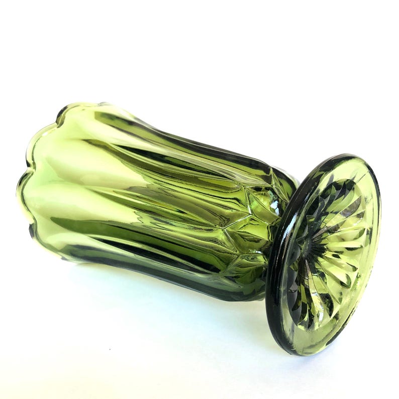 Puede incluir: Un jarr&oacute;n de vidrio verde con una parte superior acampanada y festoneada y una base redonda y estampada. El jarr&oacute;n tiene un dise&ntilde;o acanalado en el cuerpo y est&aacute; hecho de vidrio verde oliva transl&uacute;cido. La base tiene un patr&oacute;n de estrella.
