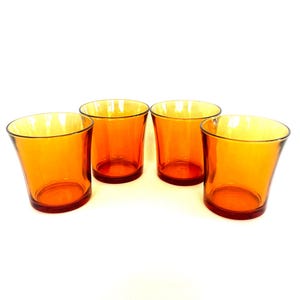 May include: Four orange-tinted glass tumblers are displayed on a white surface. The glasses have a slightly flared shape, with a thicker base and a thinner rim. The colour is a warm orange, and the glasses appear to be empty.