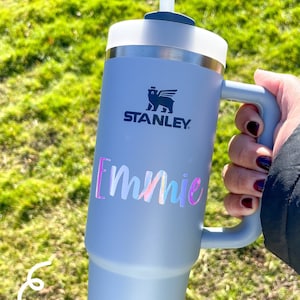 May include: A light blue Stanley tumbler with a handle and a straw. The tumbler has the name "Emmie" in a holographic font. The Stanley logo is visible. The text at the bottom says "Font: Sophia" and "Color: Opal White Holo".