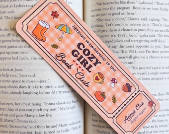 May include: A peach-colored bookmark with a gingham pattern and the text "COZY GIRL Book Club." The bookmark features illustrations of a boot, umbrella, teacup, mushroom, and pumpkin. It also has "Admit One" text.