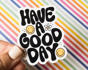 May include: A white sticker with the words "HAVE A GOOD DAY" in black bubble letters. The sticker includes smiley faces and flower graphics. The background is a colorful striped pattern.