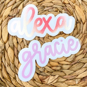 May include: Two die-cut stickers with names. The top sticker says "alexa" in a cursive font with a gradient of pink and gold. The bottom sticker says "gracie" in a pink cursive font. Both stickers have a white border.