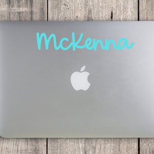 May include: A silver laptop with the Apple logo, featuring the name "McKenna" in turquoise script across the top. The laptop is set against a weathered wooden background.