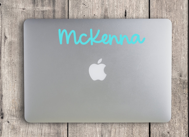 May include: A silver laptop with the Apple logo, featuring the name "McKenna" in turquoise script across the top. The laptop is set against a weathered wooden background.
