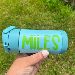 May include: A light blue water bottle with the name "MILES" in bright green letters. The bottle has a matching light blue lid and a handle. The background is green grass.